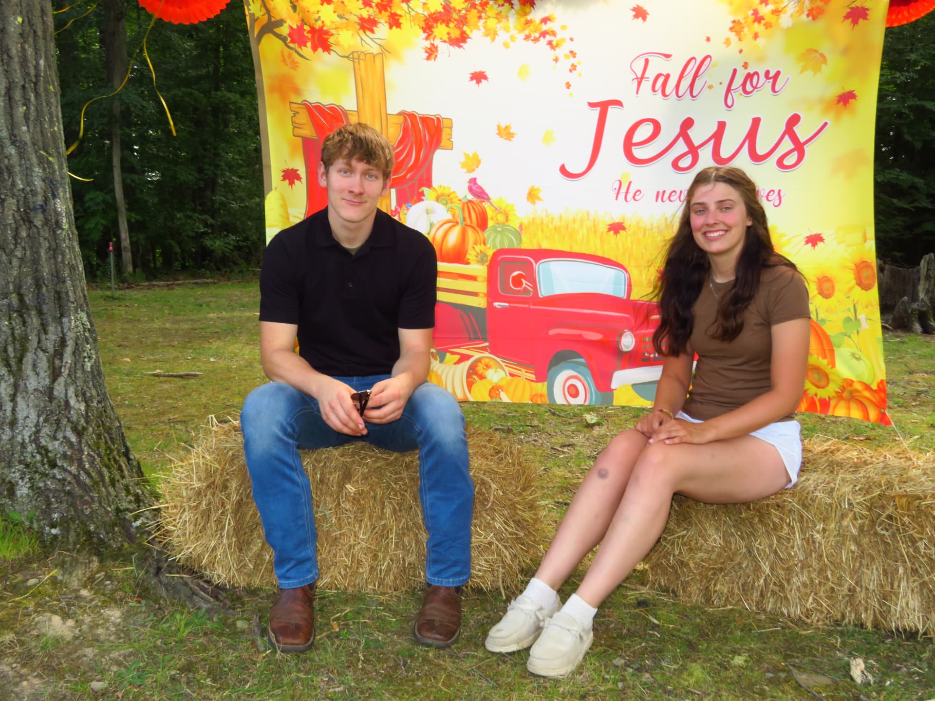 Two young adults posing on hay bales in front of the Fall for Jesus backdrop